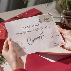 May include: A greeting card held by a person, with the text "Sometimes, One Song Says It All Curious!" and "To Julia". The card is light-colored with a floral design and is placed over a red envelope.