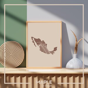 May include: A brown outline of Mexico with a stylized image of a Mayan pyramid in the center. The image is framed in a light wood frame and is displayed on a shelf with a woven basket and a vase with dried grass.