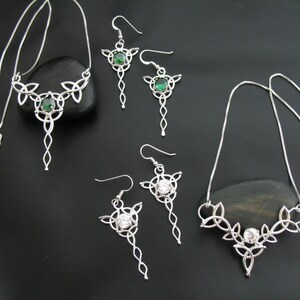 May include: A silver-toned jewelry set featuring Celtic knot designs. The set includes two necklaces and two pairs of earrings. The earrings and one necklace have green gemstones, while the other necklace and earrings have clear gemstones. The jewelry is displayed on a black surface.