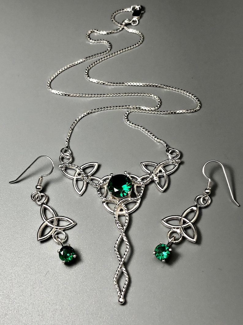 May include: A silver Celtic knot necklace and earrings set with green gemstones. The necklace features a pendant with a twisted rope design and three interconnected knots. The earrings are drop earrings with a single knot and a green gemstone.