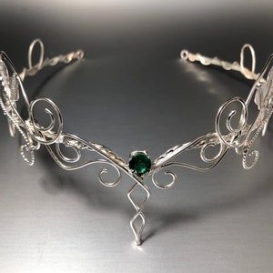 May include: Silver wire crown featuring a green gemstone at its centre. The crown has a delicate, intricate design with swirling patterns.