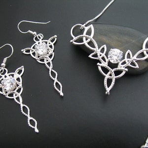 May include: A silver-toned jewelry set featuring a necklace and a pair of earrings. The necklace and earrings have a Celtic knot design with a central clear gemstone. The earrings have a long, twisted chain. The jewelry is displayed on a dark surface.