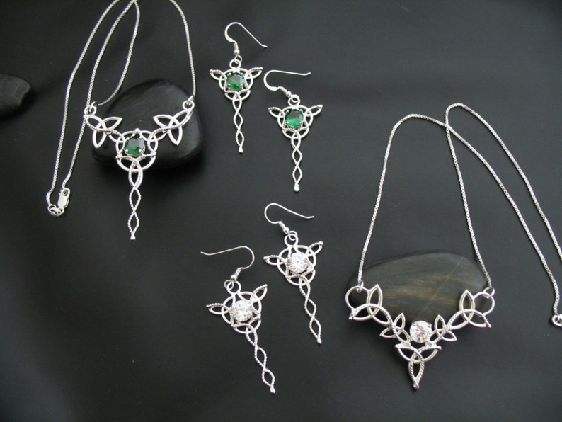 May include: A silver-toned jewelry set featuring Celtic knot designs. The set includes two necklaces and two pairs of earrings. The earrings and one necklace have green gemstones, while the other necklace and earrings have clear gemstones. The jewelry is displayed on a black surface.