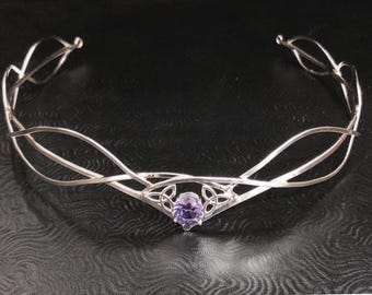 May include: A silver-toned metal circlet featuring a delicate, flowing wire design that curves around the head. The center of the piece is adorned with a single, round, faceted purple gemstone set within a decorative triquetra knot frame.