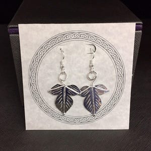 May include: A pair of silver leaf earrings with a detailed design. The earrings are hanging from a silver hook and have a small silver ring at the top.