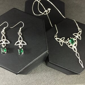 May include: A silver necklace and earrings set with a Celtic knot design and green gemstones. The necklace has a long pendant with a twisted chain.