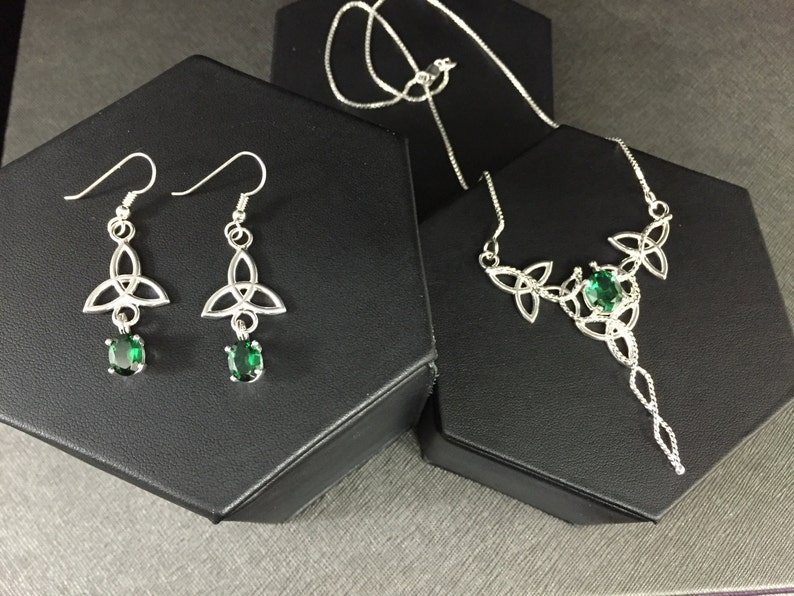 May include: A silver necklace and earrings set with a Celtic knot design and green gemstones. The necklace has a long pendant with a twisted chain.