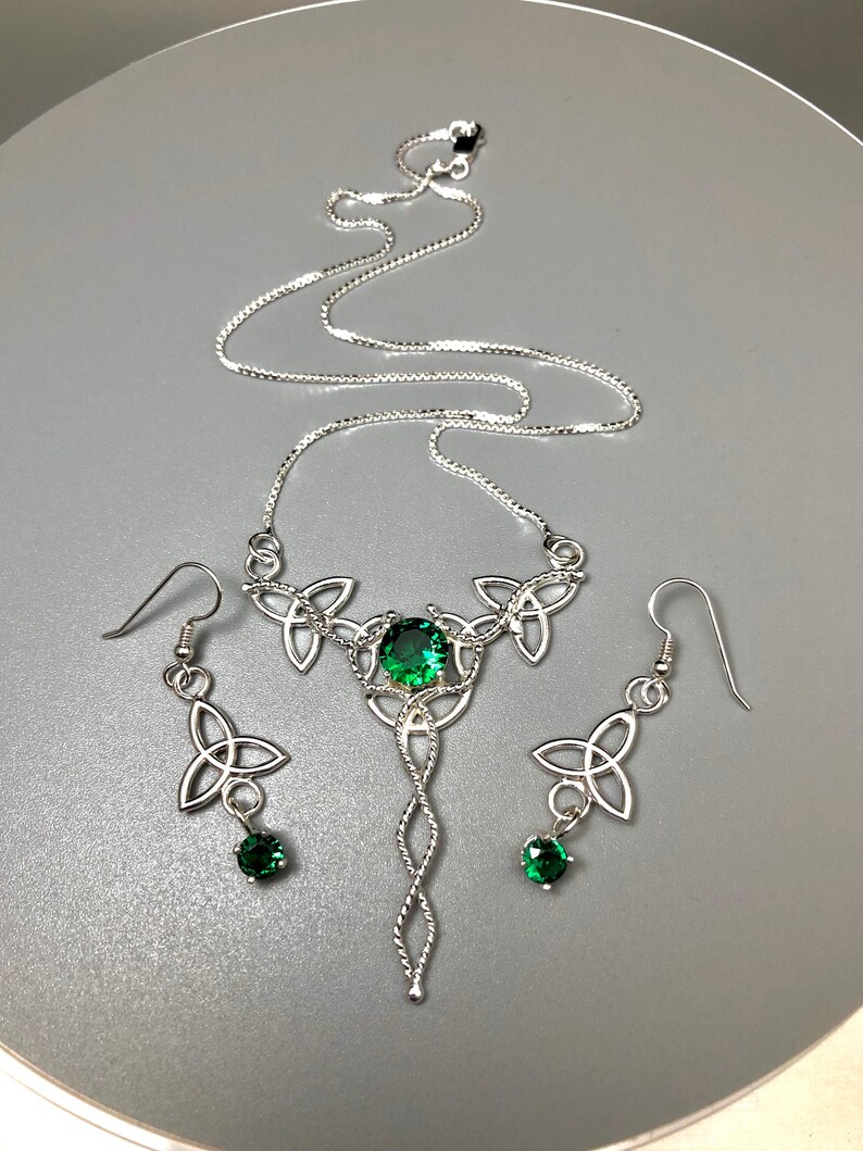 May include: A silver Celtic knot necklace and earrings set with green gemstones. The necklace features a pendant with a large green gemstone in the center and smaller green gemstones on the earrings.