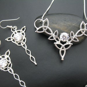 May include: A silver-toned jewelry set featuring a necklace and a pair of earrings. The necklace and earrings have a Celtic knot design with a central clear gemstone. The earrings have a long, dangling chain. The jewelry is displayed on a dark surface.
