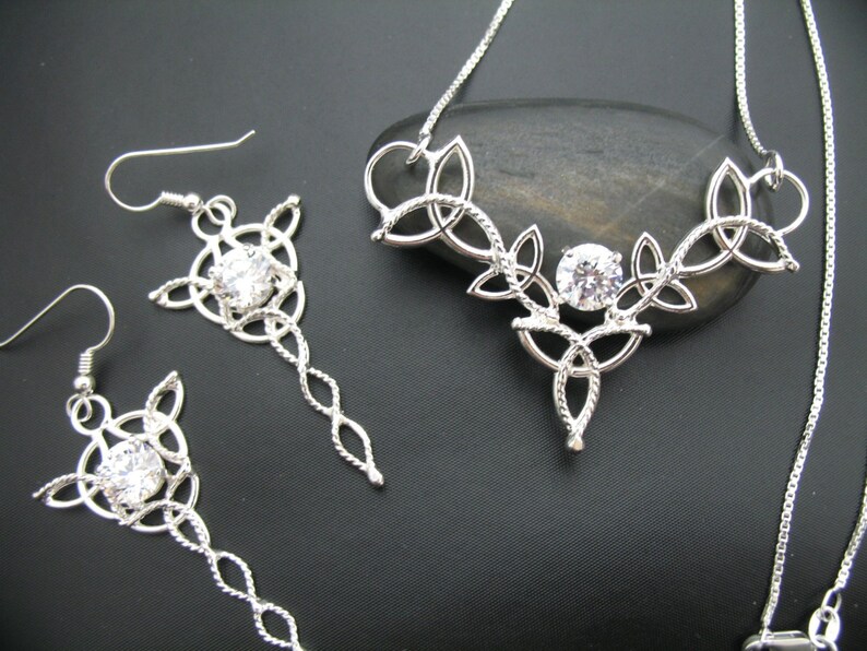 May include: A silver-toned jewelry set featuring a necklace and a pair of earrings. The necklace and earrings have a Celtic knot design with a central clear gemstone. The earrings have a long, dangling chain. The jewelry is displayed on a dark surface.