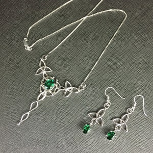May include: A silver necklace and earrings set with a Celtic knot design and green gemstones. The necklace features a long pendant with a single larger gemstone.