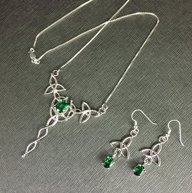 May include: A silver necklace and earrings set with a Celtic knot design and green gemstones. The necklace features a long pendant with a single larger gemstone.