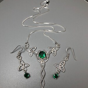 May include: A silver Celtic knot necklace and earrings set with green gemstones. The necklace features a long pendant with a twisted rope design and three smaller knots at the top. The earrings are simple drop earrings with a single knot and a green gemstone.