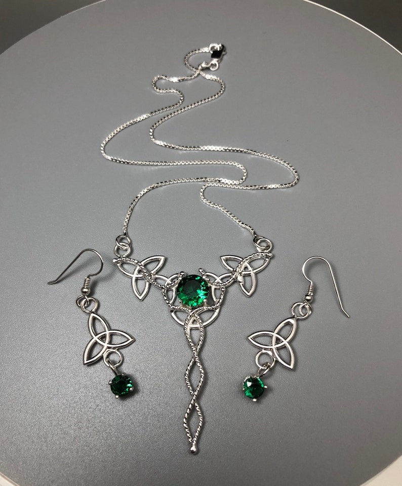 May include: A silver Celtic knot necklace and earrings set with green gemstones. The necklace features a long pendant with a twisted rope design and three smaller knots at the top. The earrings are simple drop earrings with a single knot and a green gemstone.