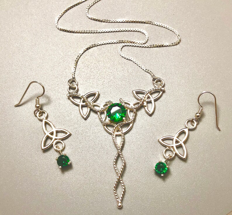 May include: A silver Celtic knot necklace and earrings set with green gemstone accents. The necklace features a pendant with a long, twisted chain.