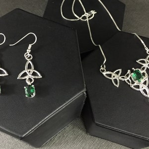 May include: A silver necklace and earrings set with a Celtic knot design and green gemstones. The necklace has a long pendant with a larger gemstone.