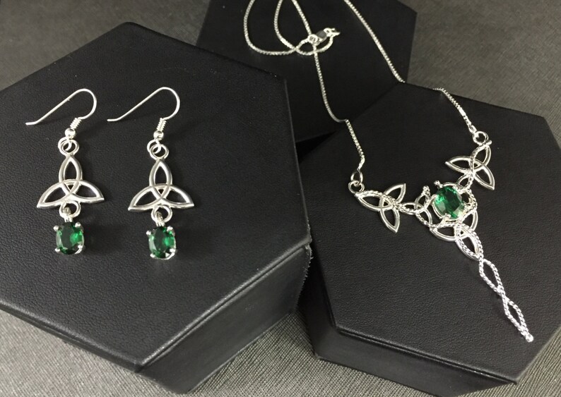 May include: A silver necklace and earrings set with a Celtic knot design and green gemstones. The necklace has a long pendant with a larger gemstone.