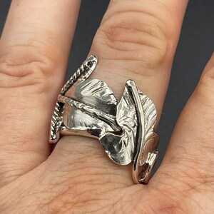 May include: A silver-toned ring featuring a textured, leaf-shaped design with a prominent central vein and a twisted rope-like detail along one side. The metal surface has a polished, reflective finish with organic, curved contours.