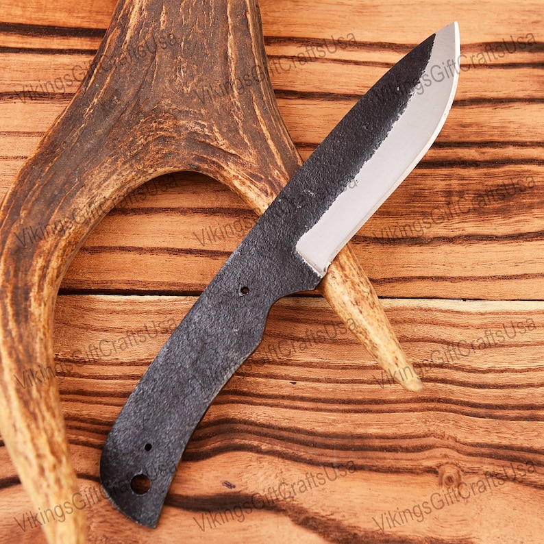 May include: A black, hand-forged knife with a slightly curved blade and a wooden handle. The knife is resting on a piece of antler.