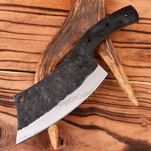 May include: A black cleaver with a curved blade and a wooden handle. The blade has a hammered finish and a small hole near the handle. The cleaver is resting on a piece of antler.
