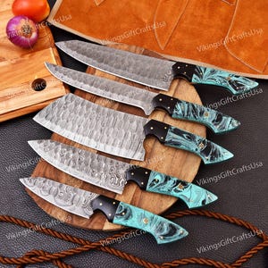 May include: Set of six kitchen knives with turquoise and black handles. The knives have a Damascus steel blade pattern and are arranged on a wooden cutting board.