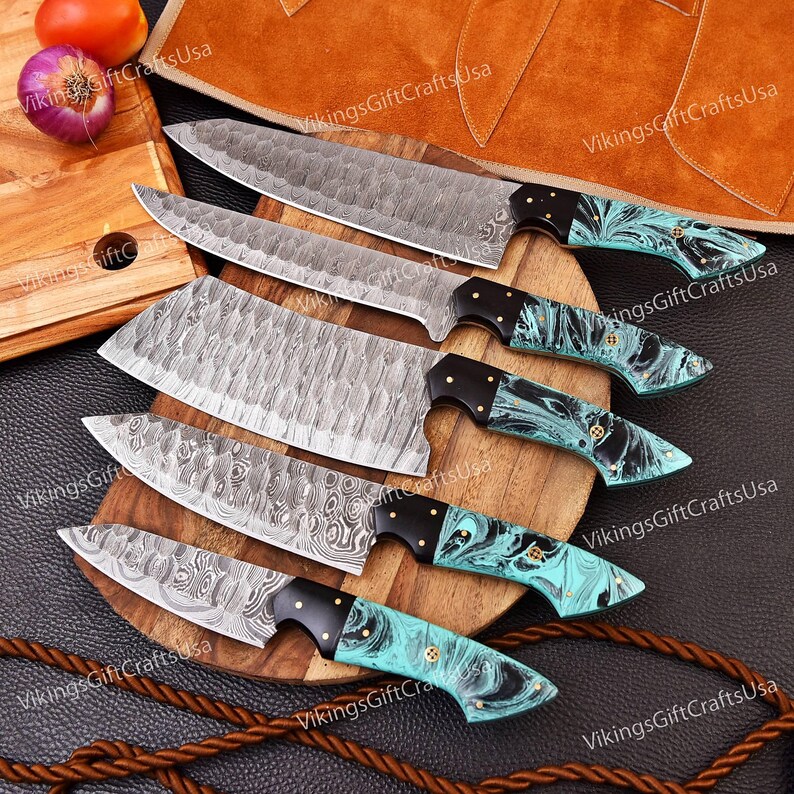 May include: Set of six kitchen knives with turquoise and black handles. The knives have a Damascus steel blade pattern and are arranged on a wooden cutting board.