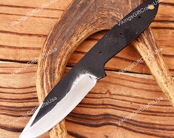 May include: A black metal knife blade with a polished silver edge. The knife has a pointed tip and a handle with three holes. The blade is set against a wooden background with a deer antler.
