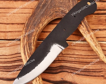 May include: A black knife blank with a slightly curved blade and a rounded tip. The knife blank is resting on a piece of antler and a wooden surface.