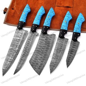 May include: Set of five kitchen knives with blue and white patterned handles. The knives have a Damascus steel blade and are displayed on a brown leather roll.