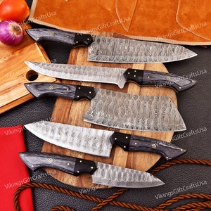 May include: Set of five kitchen knives with black wood handles and Damascus steel blades. The knives are arranged on a wooden cutting board with a brown leather background.