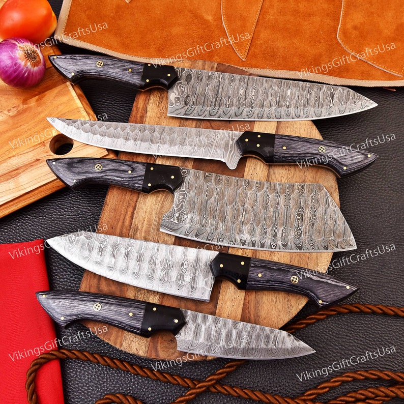 May include: Set of five kitchen knives with black wood handles and Damascus steel blades. The knives are arranged on a wooden cutting board with a brown leather background.