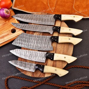May include: Set of five kitchen knives with patterned blades and bone-colored handles. The knives are arranged on a wooden cutting board with a brown leather background.
