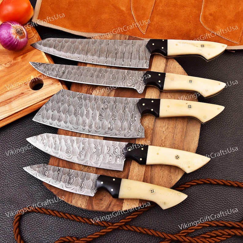 May include: Set of five kitchen knives with patterned blades and bone-colored handles. The knives are arranged on a wooden cutting board with a brown leather background.