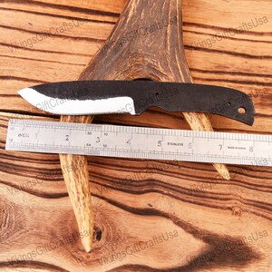 May include: A black and white knife with a rounded tip and a hole near the handle. The knife is laying on a wooden surface with a ruler and a piece of antler.