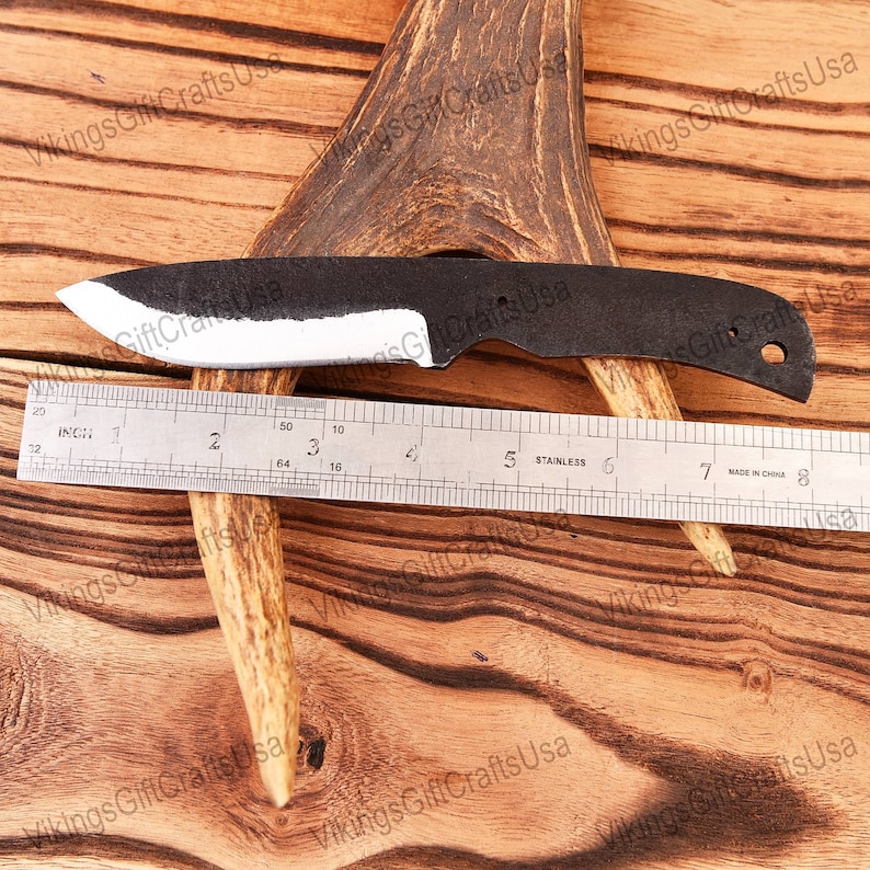 May include: A black and white knife with a rounded tip and a hole near the handle. The knife is laying on a wooden surface with a ruler and a piece of antler.