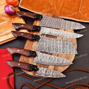 May include: Set of six kitchen knives with wooden handles and Damascus steel blades. The knives are arranged on a wooden cutting board and a brown leather surface.