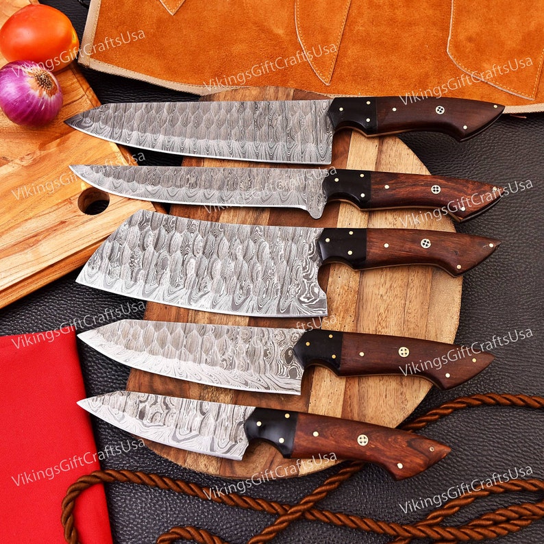 May include: Set of five kitchen knives with Damascus steel blades and wooden handles. The knives are arranged on a wooden cutting board with a brown leather background.