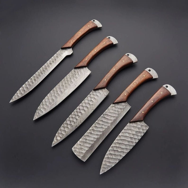 May include: Set of five kitchen knives with wooden handles and a patterned steel blade. The knives are arranged in a row on a black surface.
