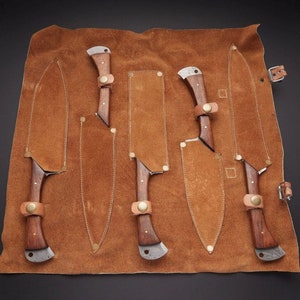 May include: A set of five knives with wooden handles and leather sheaths, arranged in a brown leather roll.
