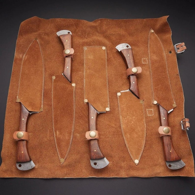 May include: A set of five knives with wooden handles and leather sheaths, arranged in a brown leather roll.
