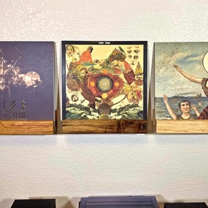 May include: Three rectangular wooden wall-mounted shelves holding vinyl record covers. The left cover is dark with a tree graphic, the center cover features a colorful abstract illustration with the text Fleet Foxes, and the right cover shows a surreal painting.