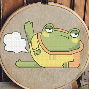 May include: Cross-stitch embroidery in a wooden hoop shows a cartoon frog in a yellow outfit with a white speech bubble. The frog is green and yellow with a grumpy expression. The background includes a stack of books and a floral painting.