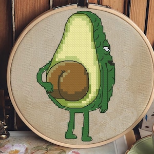 Puede incluir: Un bastidor de bordado de punto de cruz con un personaje de aguacate de dibujos animados. El aguacate es verde y amarillo con un hueso marrón, y tiene brazos, piernas y una expresión de mal humor. El bordado está sobre una tela beige.