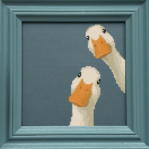 May include: A framed cross-stitch artwork featuring two white ducks with orange beaks peeking out from behind a blue background. The frame is a light teal color. The artwork is square in shape.