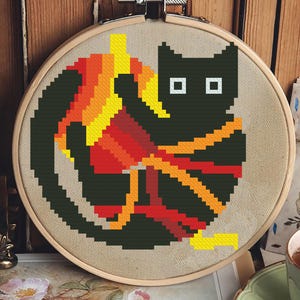 May include: Cross-stitch embroidery of a black cat curled around a ball of yarn. The yarn is a mix of red, orange, and yellow. The embroidery is set in a wooden hoop, displayed on a table with a painting and a cup of tea.