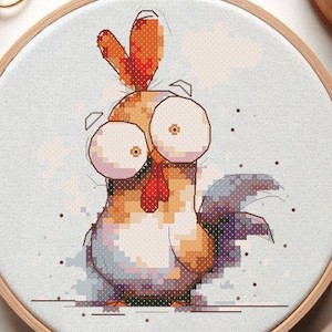 Puede incluir: Un aro de bordado de punto de cruz con un fondo blanco y un gallo de dibujos animados con ojos grandes y una cresta roja. El gallo es marrón y naranja con una cola morada.