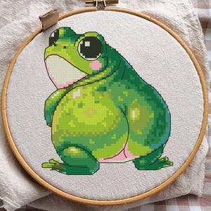 Peut inclure: Broderie au point de croix d'une grenouille verte aux grands yeux et au ventre rose, placée dans un cercle en bois. La grenouille est réalisée avec des détails pixélisés. Le fond est un tissu neutre.