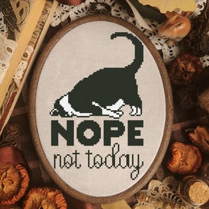 Puede incluir: Un punto de cruz ovalado enmarcado, que representa un gato blanco y negro con la espalda arqueada. El texto "NOPE not today" está bordado debajo. El marco es de color marrón oscuro.