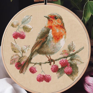 Puede incluir: Bordado de punto de cruz terminado de un petirrojo posado en una rama con bayas rojas. El ave tiene el pecho naranja y está rodeada de follaje verde y beige. El bordado está en un bastidor de madera.
