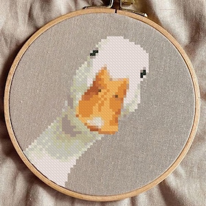 Op de afbeelding: Een kruissteekborduurwerk van een ganzenkop, met witte en oranje gepixelde details. Het kunstwerk is geplaatst in een houten borduurring, tegen een neutraal gekleurde stoffen achtergrond. De gans kijkt de kijker recht aan.